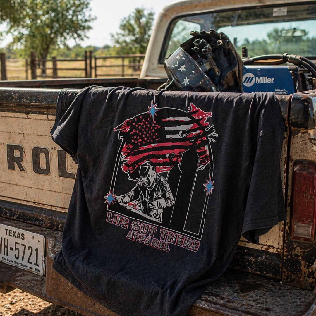 The Tack It Tee - Black by Life Out There Apparel is a unisex graphic shirt for welders, featuring a stylized U.S. flag and a welder. It's draped over an old brown Chevy truck with welding gear and Texas plate HH-5721 nearby.