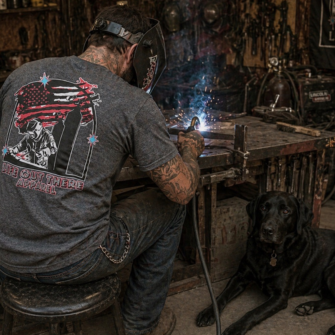 Wearing the Tack It Tee - Black Heather from Life Out There Apparel and a welding helmet, a welder works at a bench as a black labrador dog looks at the camera in a workshop filled with tools—a true haven for welders ready to tack it.