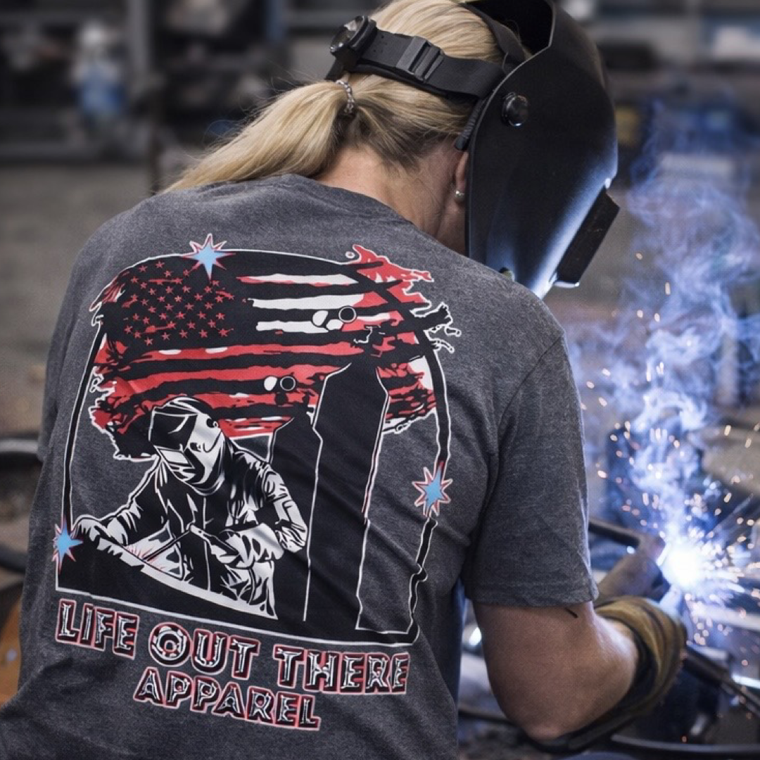 Wearing the Tack It Tee - Black Heather by Life Out There Apparel, a welder dons a mask and gloves as sparks fly. The unisex tee shows a welder, two towers, an American flag, and LIFE OUT THERE APPAREL on the back as they TACK IT with skill.