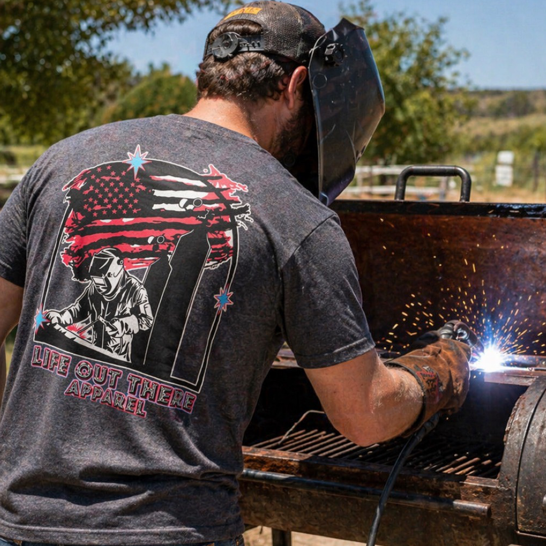 A person wearing the Tack It Tee - Black Heather by Life Out There Apparel welds outdoors, with trees and blue sky in the background. The unisex shirt displays a welder with an American flag and "Life Out There Apparel" text.