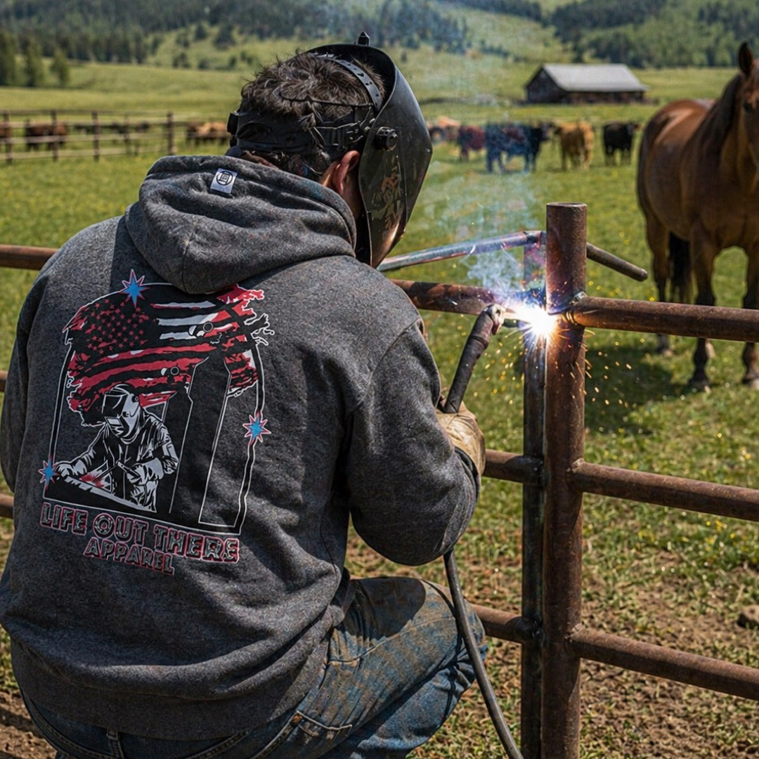 A man wearing the Life Out There Apparel Tack It Hoodie in  Black Heather with welder graphics on it plus an American flag, a welder in a helmet repairs a metal fence outdoors near horses and a barn on a grassy field.