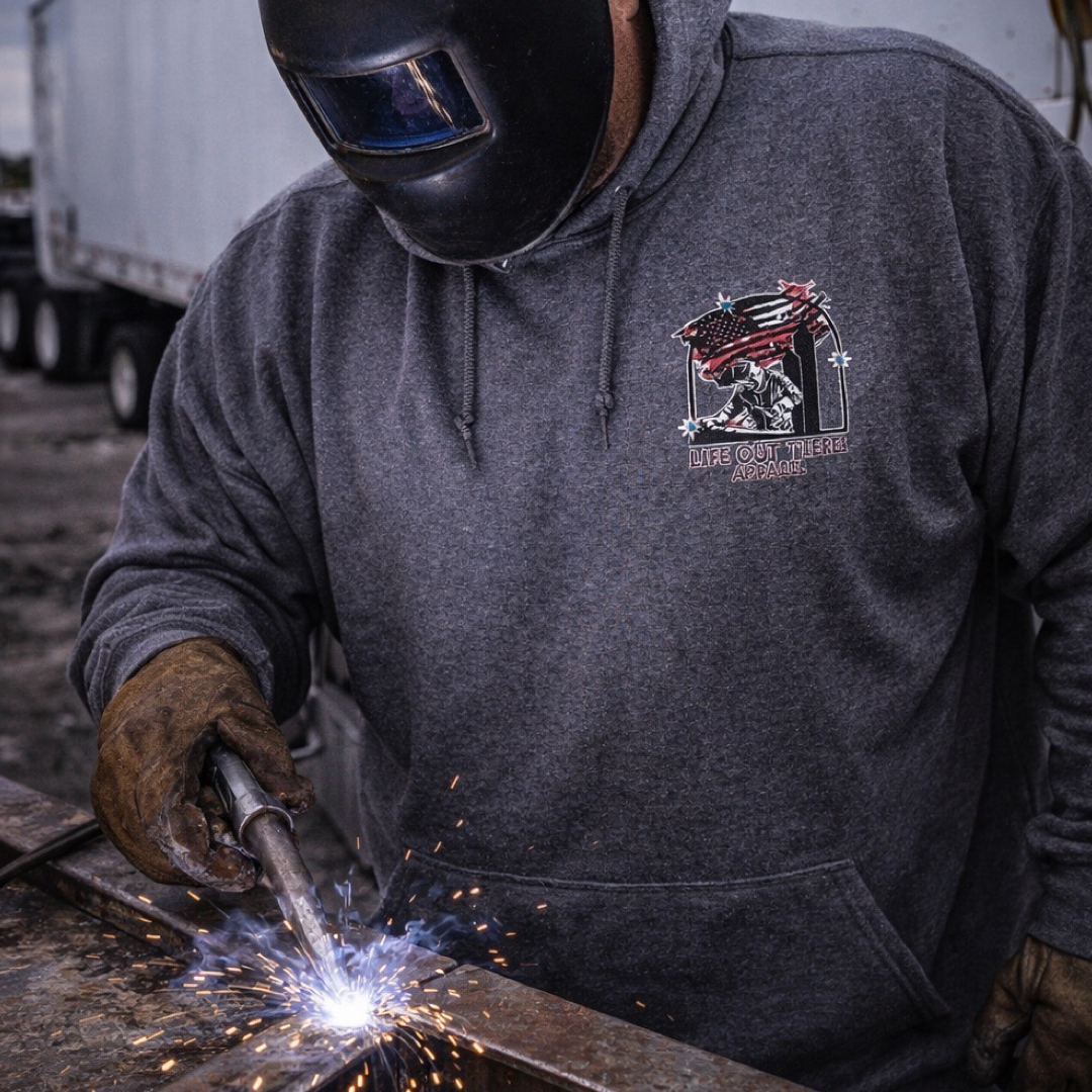 Wearing the Life Out There Apparel Tack It Hoodie in Black Heather, a welder dons a helmet and gloves, fusing metal as sparks fly. A welder and sparks on the hoodie nods to their skill, with a big truck blurred in the background.