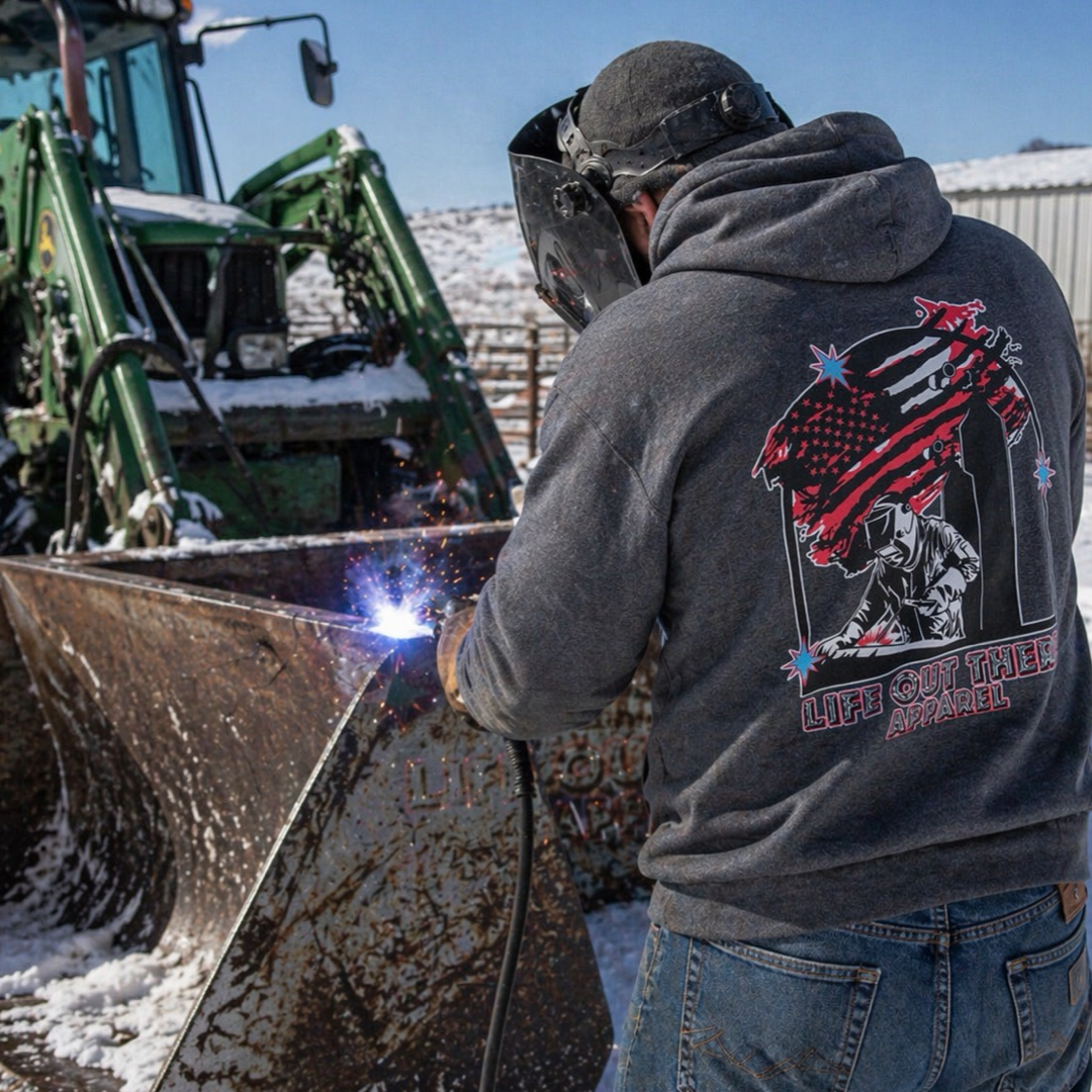 A person welds metal outdoors next to a snow-covered tractor bucket, wearing a TACK IT welding helmet and the Life Out There Apparel Tack It Hoodie in Black Heather featuring an American flag design on the back.