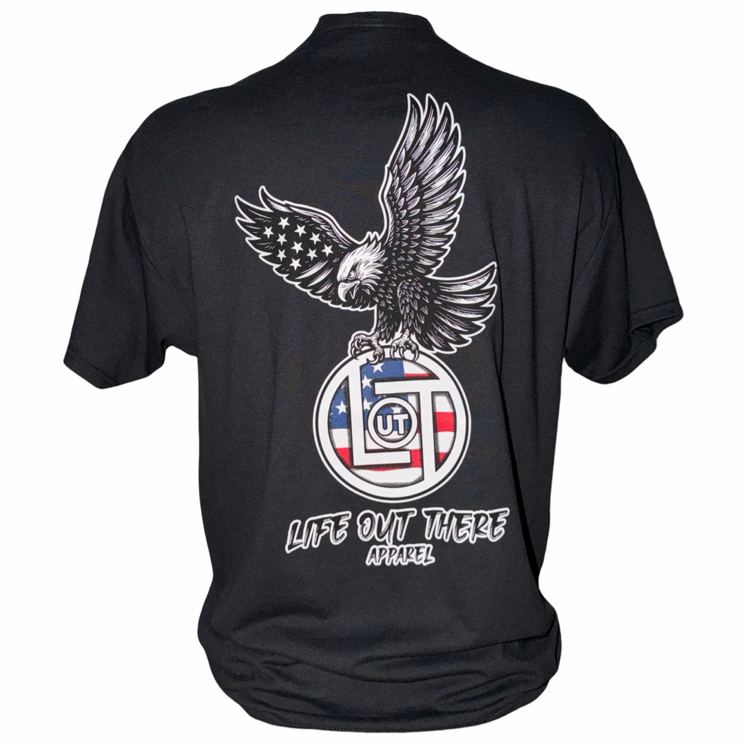 The Flight of Freedom Tee - Black by Life Out There Apparel is a tagless unisex t-shirt with a graphic of a bald eagle on a circular stars-and-stripes emblem with "OUT" lettering and the text LIFE OUT THERE APPAREL below.