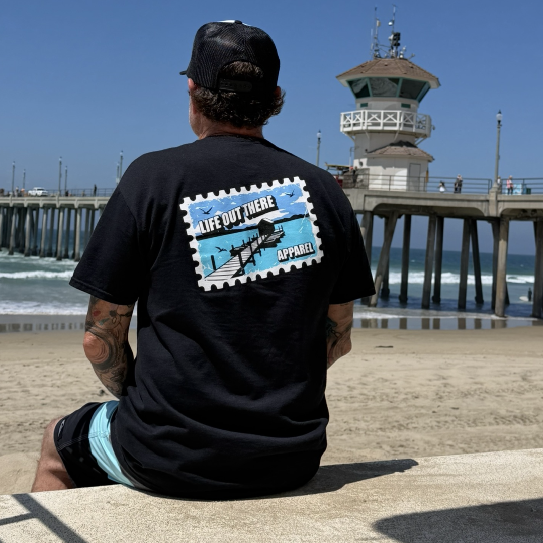 A man in a black Wish You Were Here Tee by Life Out There Apparel and a cap sits on a concrete ledge at the beach, facing a pier. The shirt’s back shows a postcard design with “Wish You Were Here” and a lifeguard tower image.