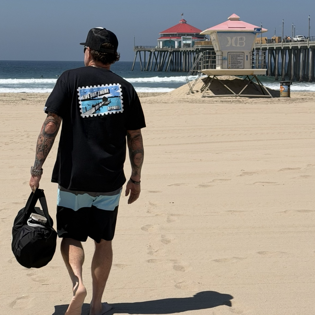A man in the Life Out There Apparel Wish You Were Here Tee - Black walks barefoot on the sandy beach, carrying a duffel bag. A pier and lifeguard tower stand in the background, evoking a classic postcard vibe.