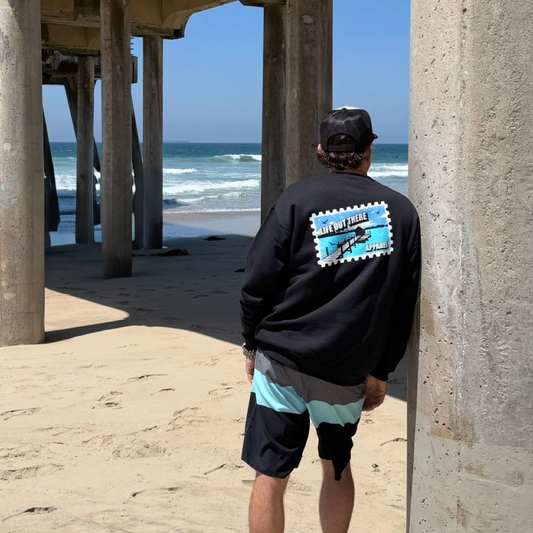 A person wearing the Wish You Were Here Crewneck Sweatshirt - Black by Life Out There Apparel leans against a pier pillar, facing the ocean on a sandy beach. The sweatshirt features a blue LIFE OUT THERE graphic.