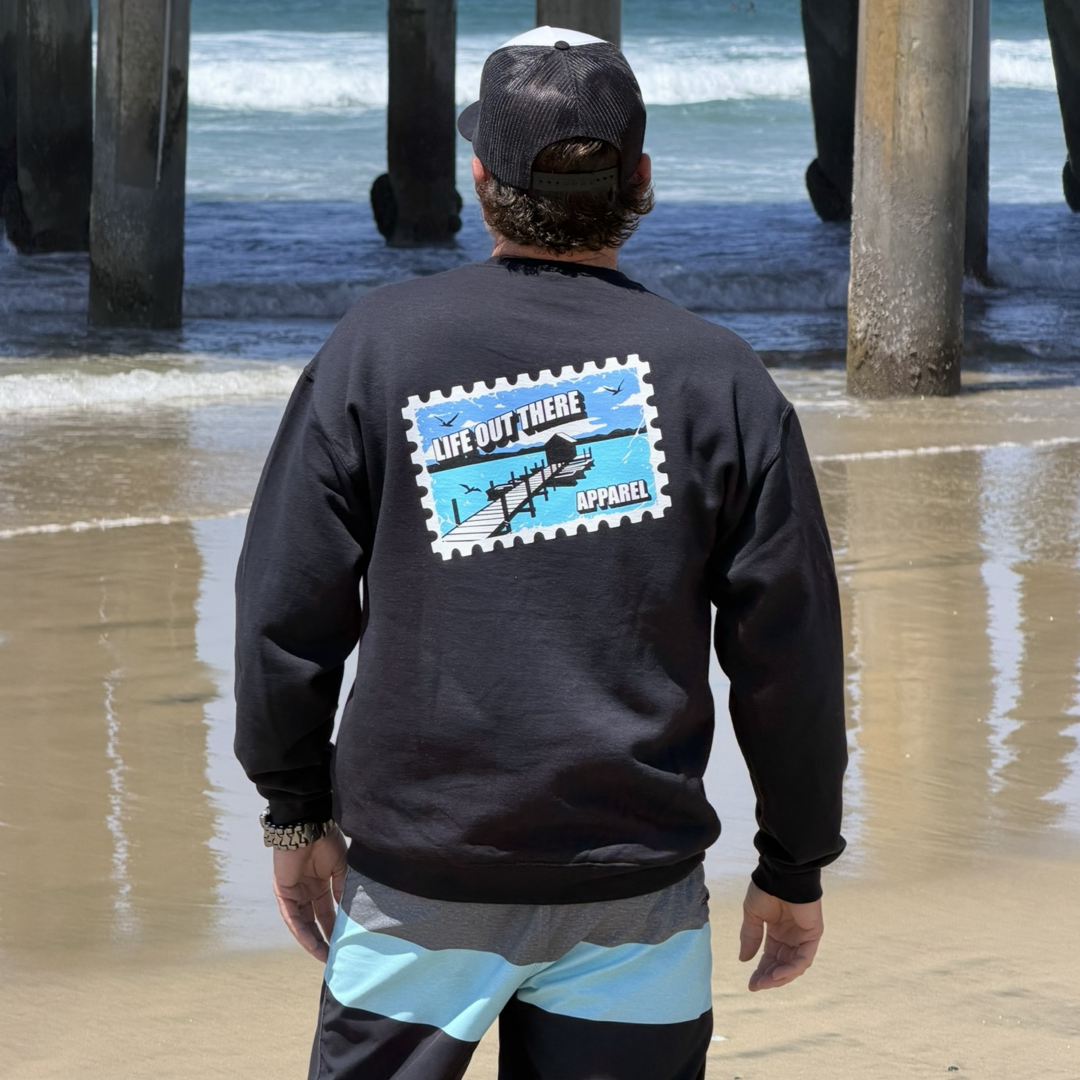 Standing on a sandy beach near a pier, someone faces the ocean in a black Wish You Were Here Crewneck Sweatshirt from Life Out There Apparel, featuring a blue and white postcard graphic on the back and paired with a black cap as waves roll in.