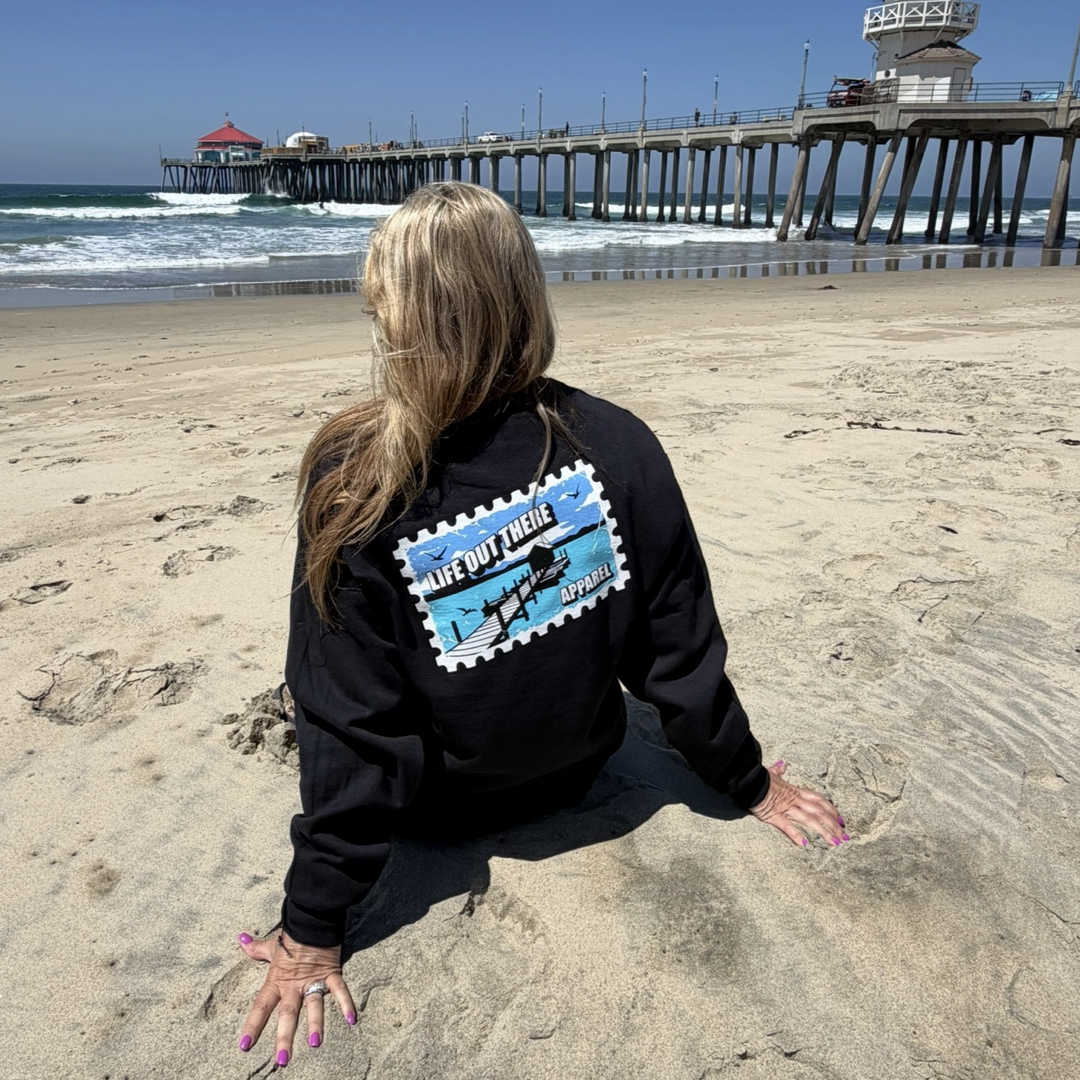 A woman with long blonde hair sits on a sandy beach facing a pier, wearing the Wish You Were Here Crewneck Sweatshirt in black by Life Out There Apparel. The ocean and blue sky stretch out in the background.