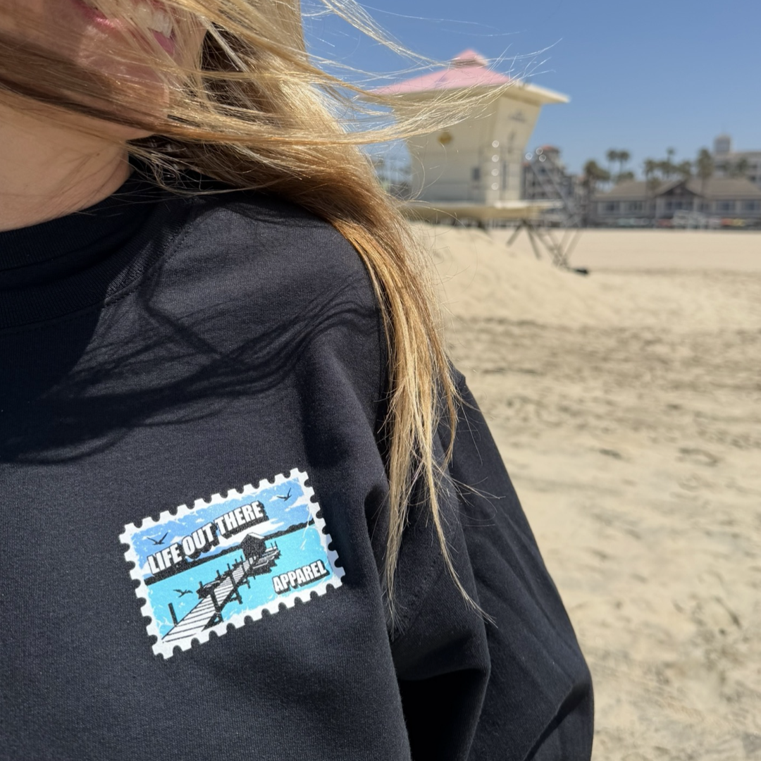 A person in a black "Wish You Were Here" crewneck sweatshirt by Life Out There Apparel stands on a sandy beach with a lifeguard tower and houses behind them, beneath a clear blue sky, giving off classic postcard vibes.