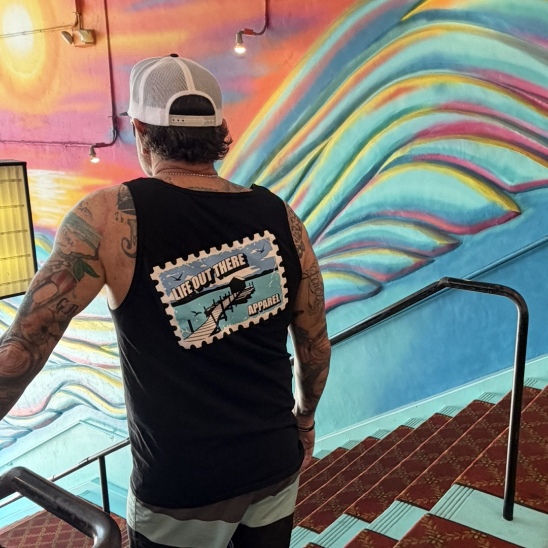 A tattooed person in a black "Wish You Were Here" tank top by Life Out There Apparel and a white cap stands on carpeted stairs, facing a mural of a sun and blue waves. The tank top features the brand’s name and a fish graphic.