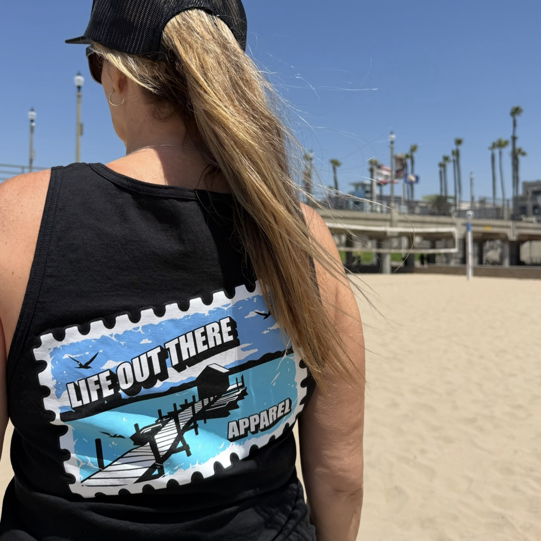 A woman with long blonde hair, wearing a black cap and the "Wish You Were Here Tank Top - Black" from Life Out There Apparel, stands on a sunny beach with palm trees and buildings in the background.