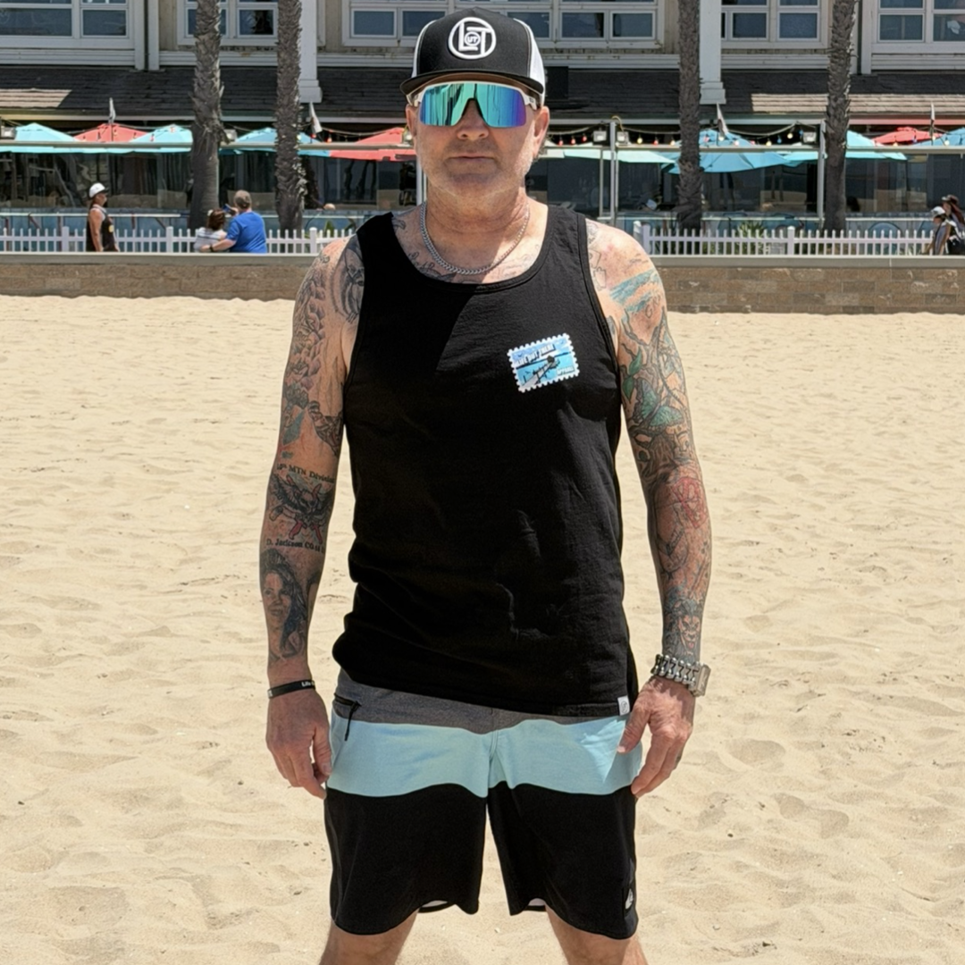 Wearing the Wish You Were Here Tank Top - Black by Life Out There Apparel, a tattooed man stands on a sandy beach with palm trees and a building behind him, showing off his ideal summer style.