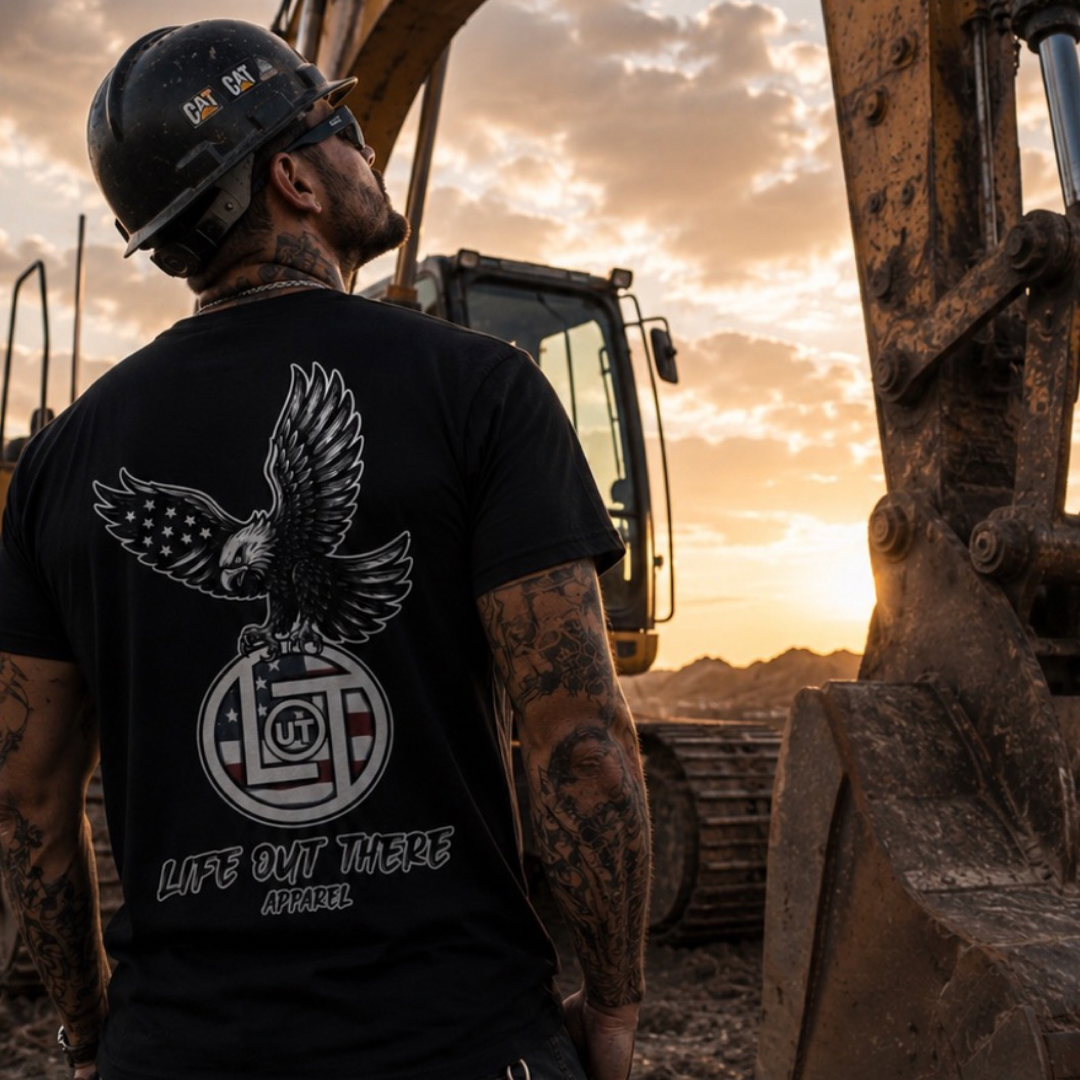 A tattooed man wears the black Flight of Freedom Tee by Life Out There Apparel at a sunset construction site, showing off its eagle and US flag design—an American symbol of the FLIGHT of FREEDOM spirit.