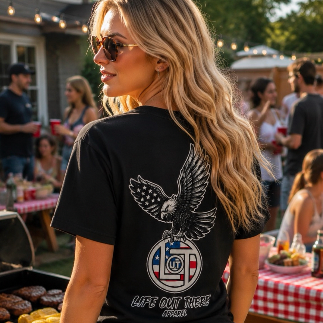 A woman with long blonde hair, sunglasses, and a Life Out There Apparel Flight of Freedom Tee - Black with an eagle and American flag stands at an outdoor barbecue, people chatting in the background at picnic tables.