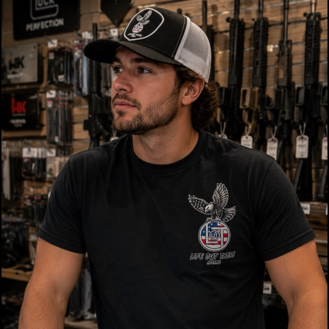 A man with a beard and long hair, wearing a black cap and a Life Out There Apparel Flight of Freedom Tee - Black that reads LIFE OUT WEST, stands in a gun store with rifles lined up on the wall behind him.