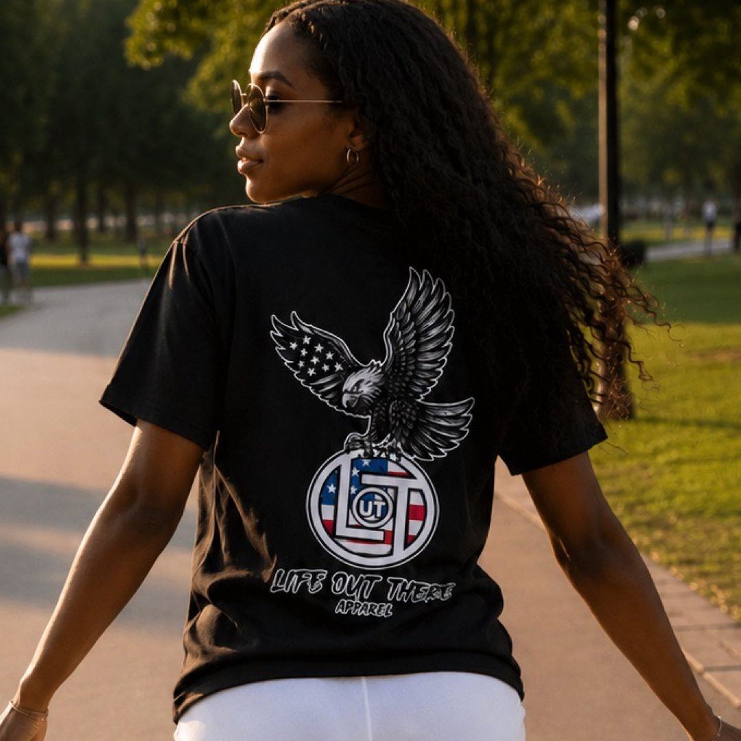 A woman with long curly hair walks outdoors wearing sunglasses and a black Flight of Freedom Tee by Life Out There Apparel, featuring an eagle holding an American flag emblem and “LIFES OUT THERE APPAREL” text on the back.