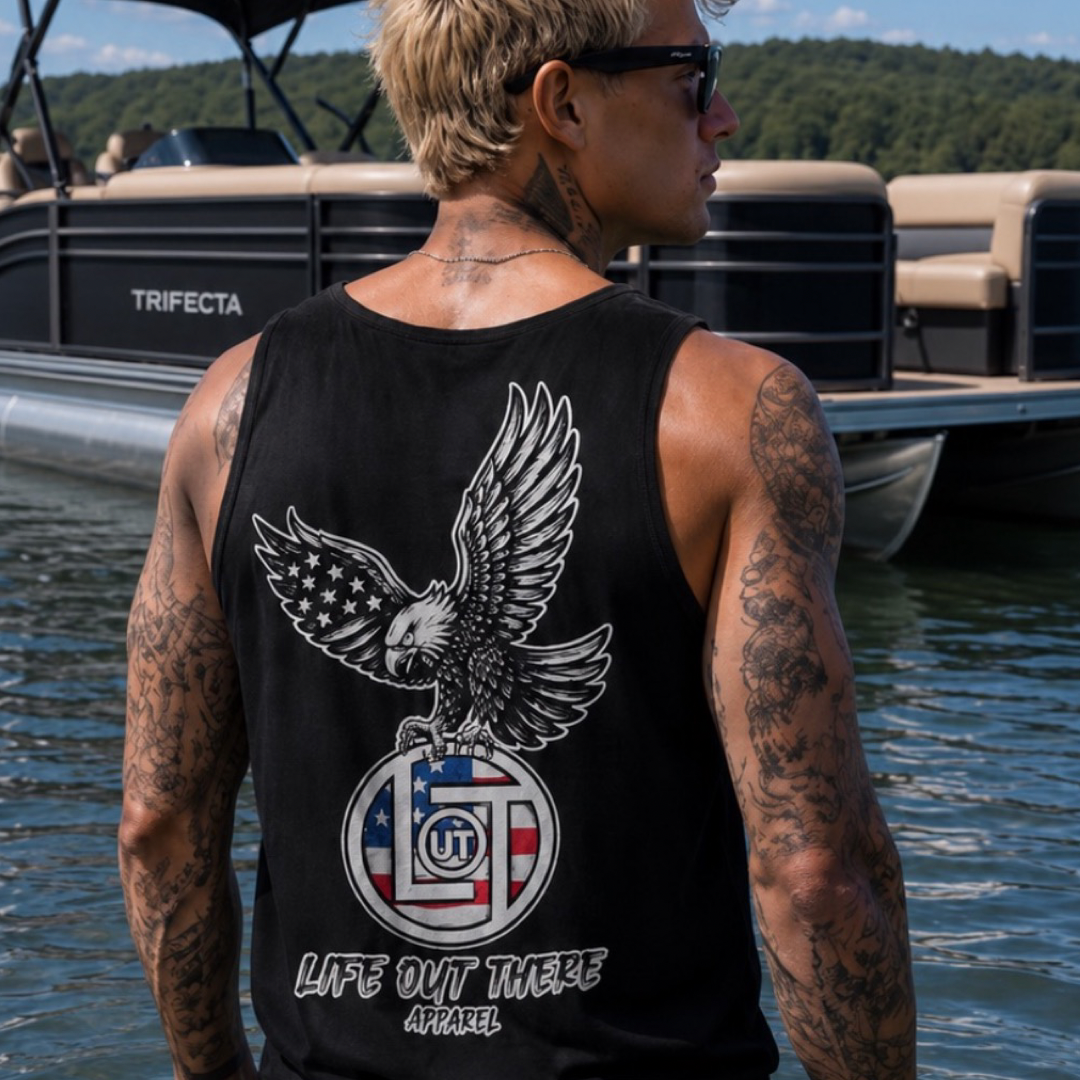 A tattooed man stands by a boat, facing away, wearing the Flight of Freedom Tank Top - Black from Life Out There Apparel. The background features water and greenery.