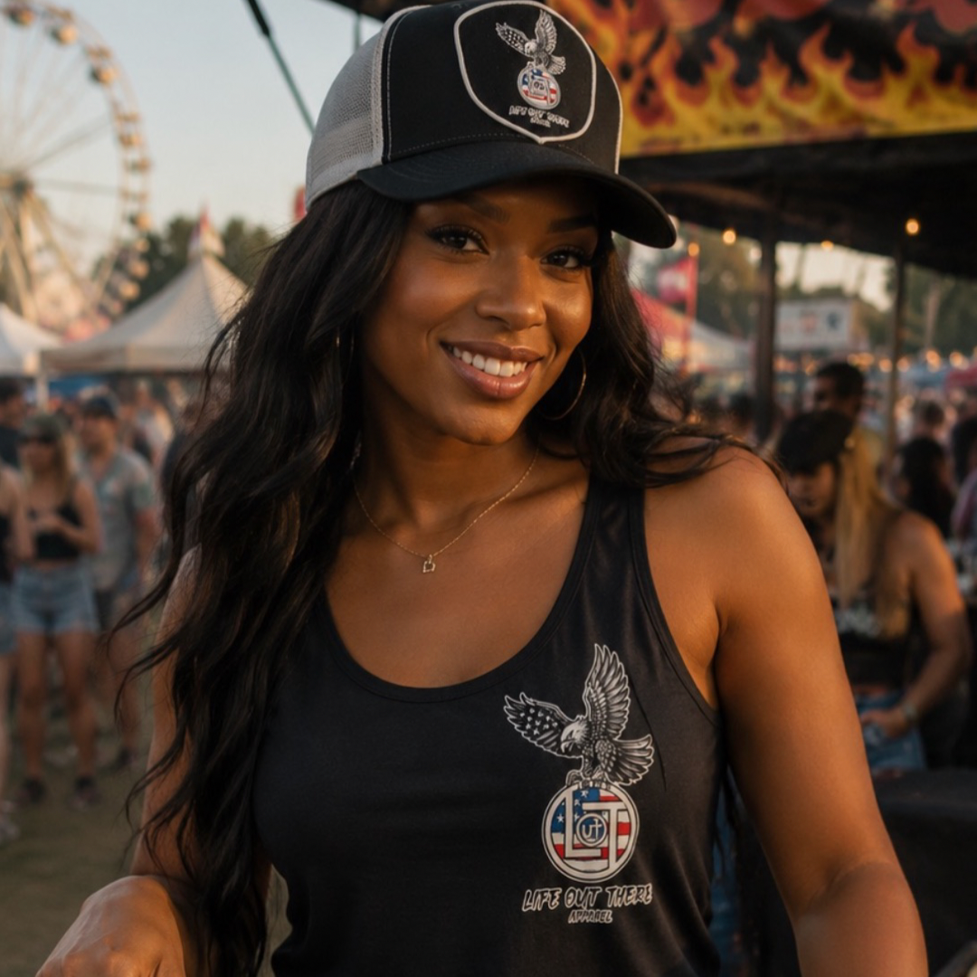 A woman in the Flight of Freedom Tank Top - Black by Life Out There Apparel smiles at an outdoor fair, with a carnival ride and people blurred in the background, capturing the vibrant spirit of summer events.