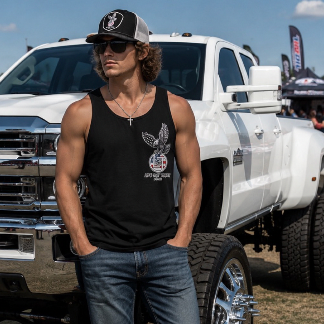 A man in jeans and sunglasses stands in front of a large white pickup at an outdoor event, proudly wearing the Life Out There Apparel Flight of Freedom Tank Top in black, with other vehicles and people seen in the background.