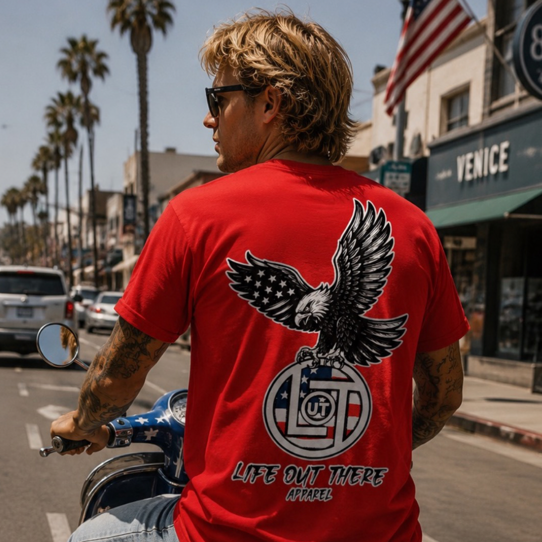 A blonde, tattooed man rides a scooter on a palm-lined street, wearing the red Flight of Freedom Tee by Life Out There Apparel with American flag and eagle motifs. An American flag waves ahead.