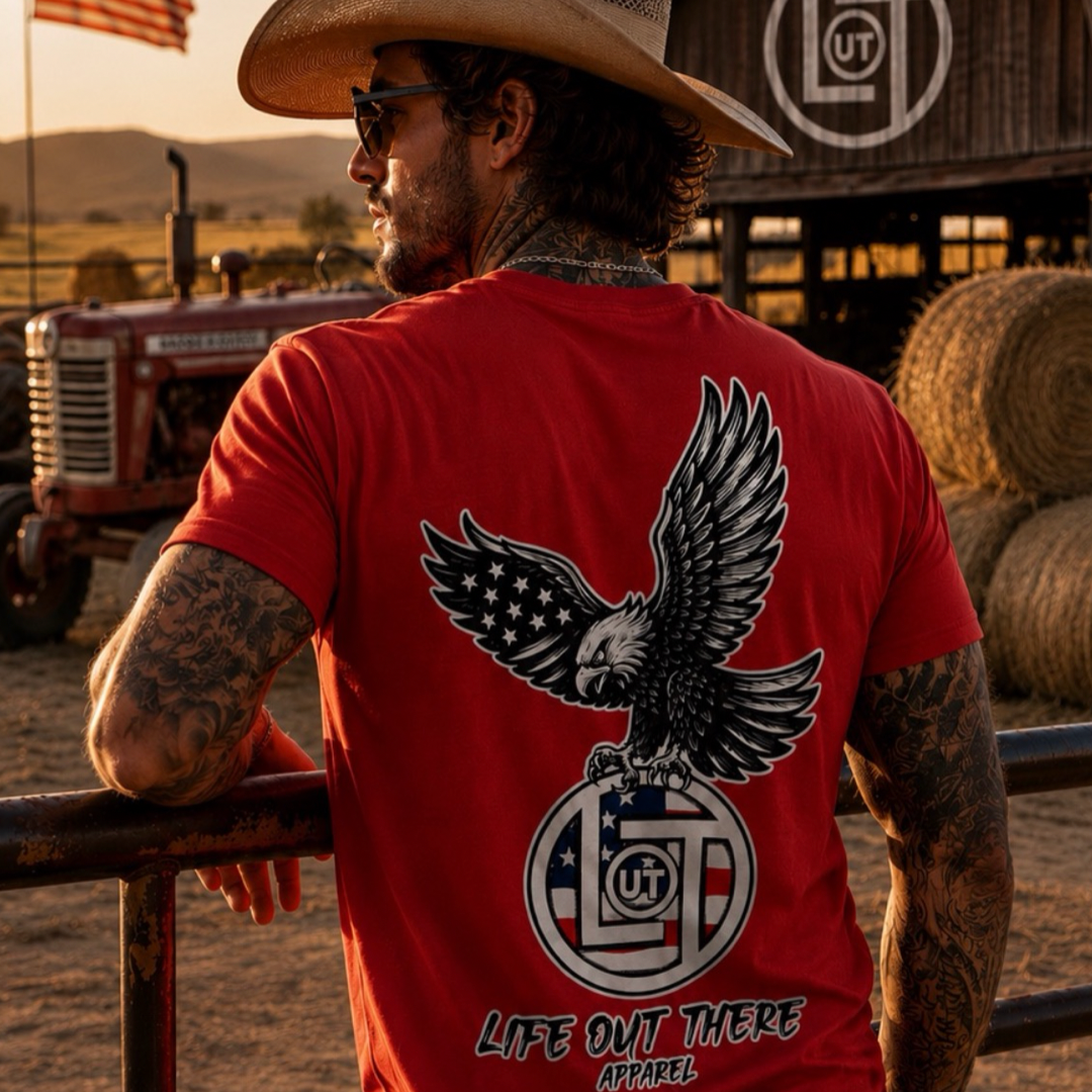 A man in a cowboy hat stands by a farm fence, wearing the red Flight of Freedom Tee from Life Out There Apparel, featuring an eagle and American flag design. Hay bales, barn, and tractor set the rural scene—true American spirit.