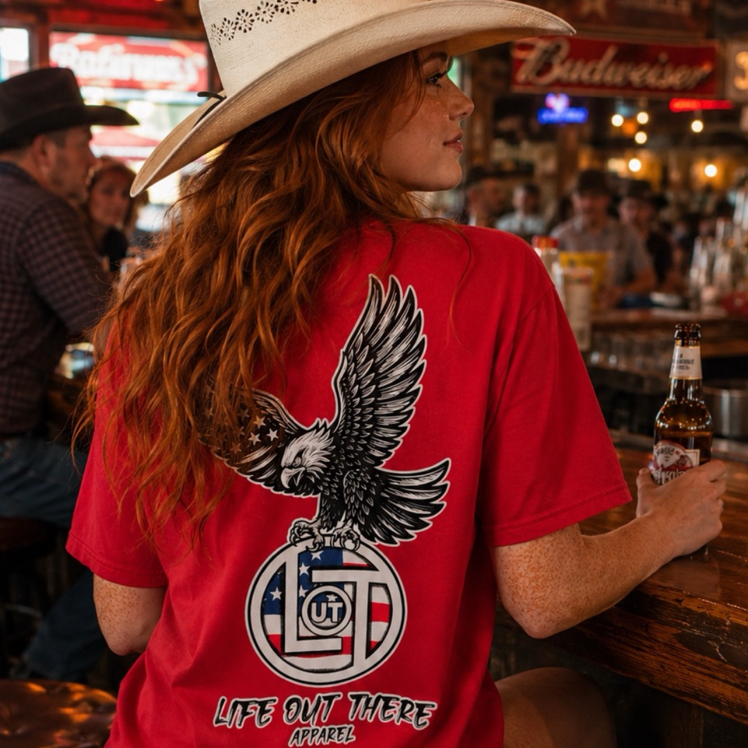 A woman in a cowboy hat and the red Flight of Freedom Tee by Life Out There Apparel sits at a bar holding a bottle, with rustic decor, other patrons, and a Budweiser sign visible in the background.