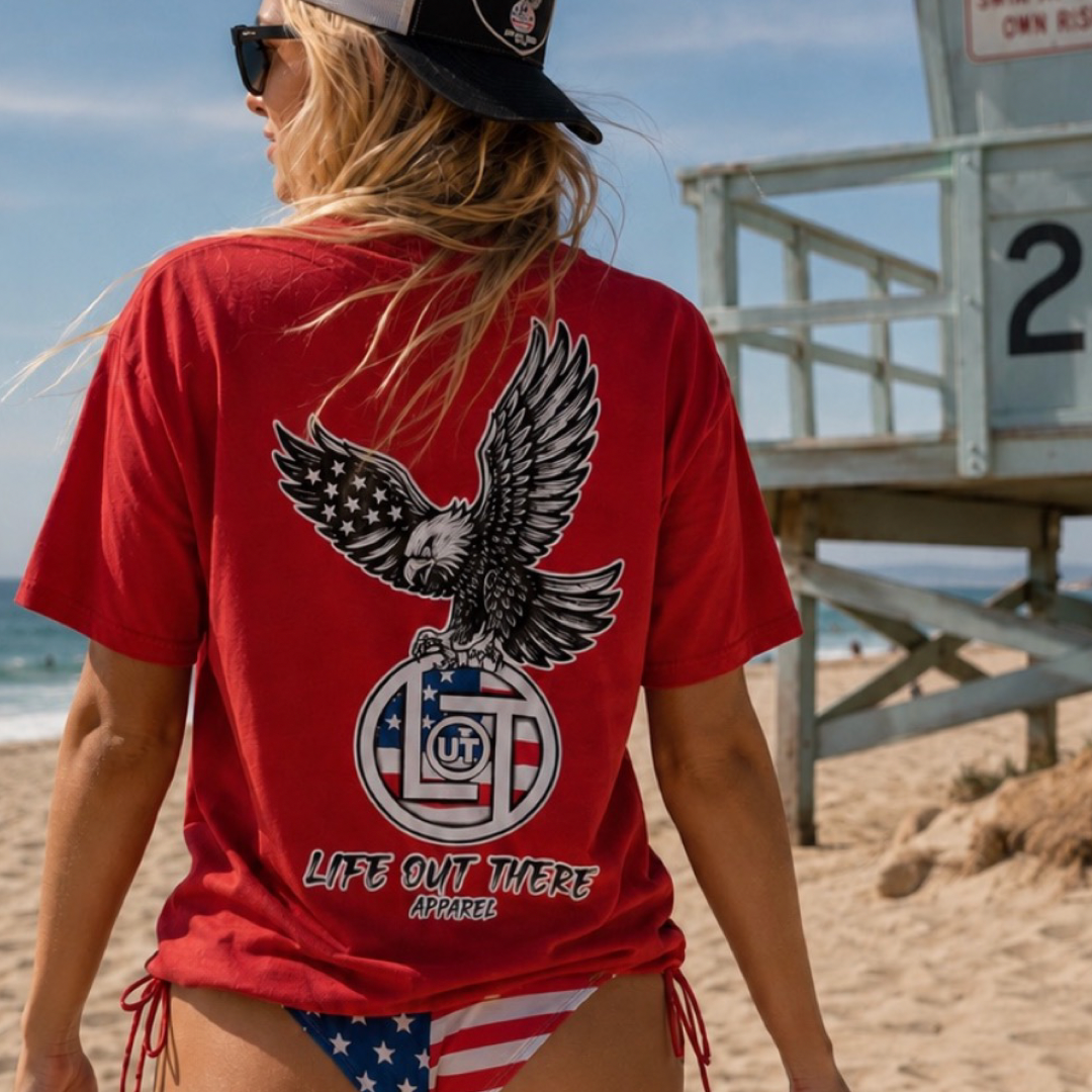 A woman on a sandy beach wears the red Flight of Freedom Tee by Life Out There Apparel with an eagle graphic, stars and stripes bikini bottoms, sunglasses, and a black cap. A lifeguard tower is in the background.