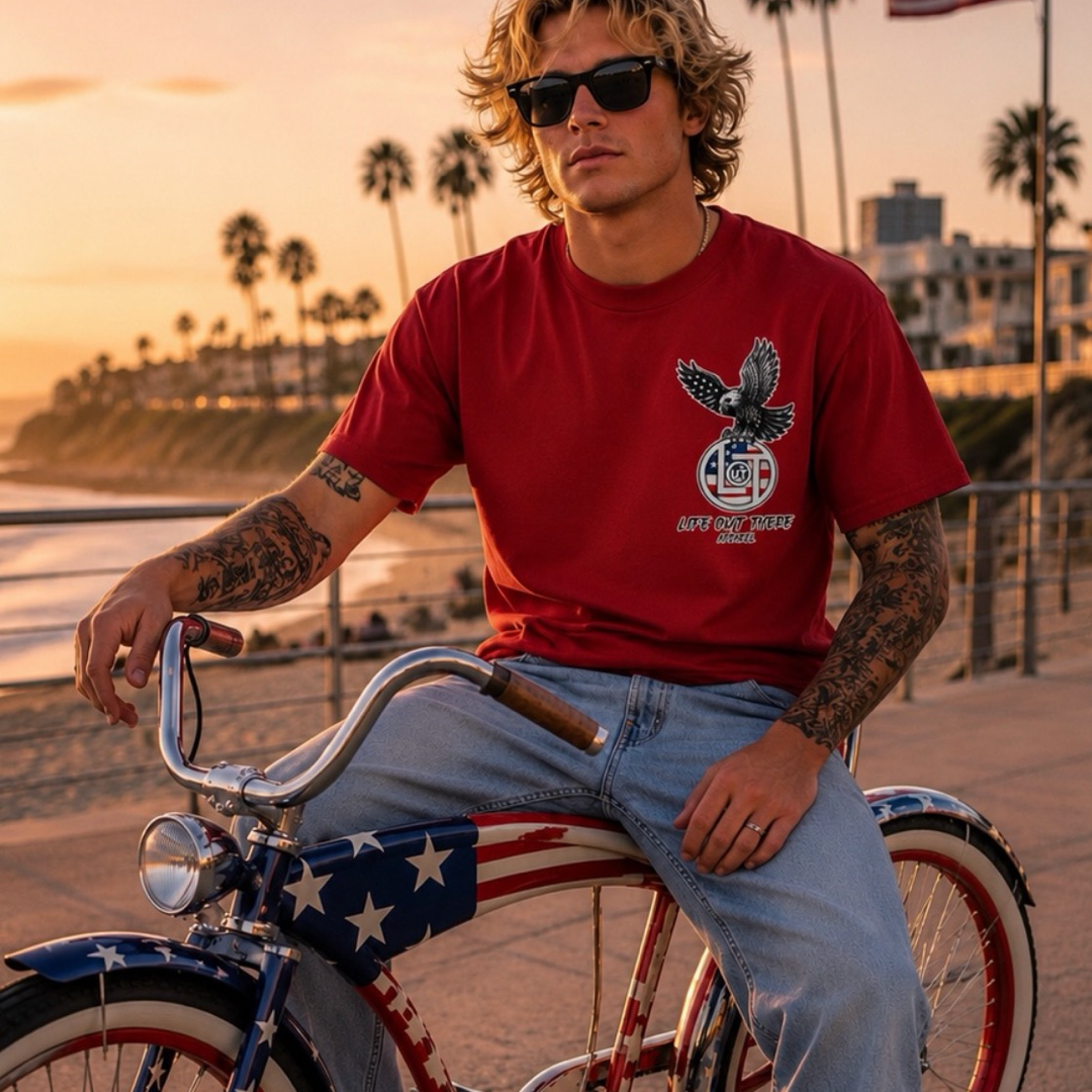 A tattooed man in sunglasses sits on a star-spangled bicycle by the beach at sunset, wearing the red Flight of Freedom Tee from Life Out There Apparel, with palm trees and an American flag behind him.