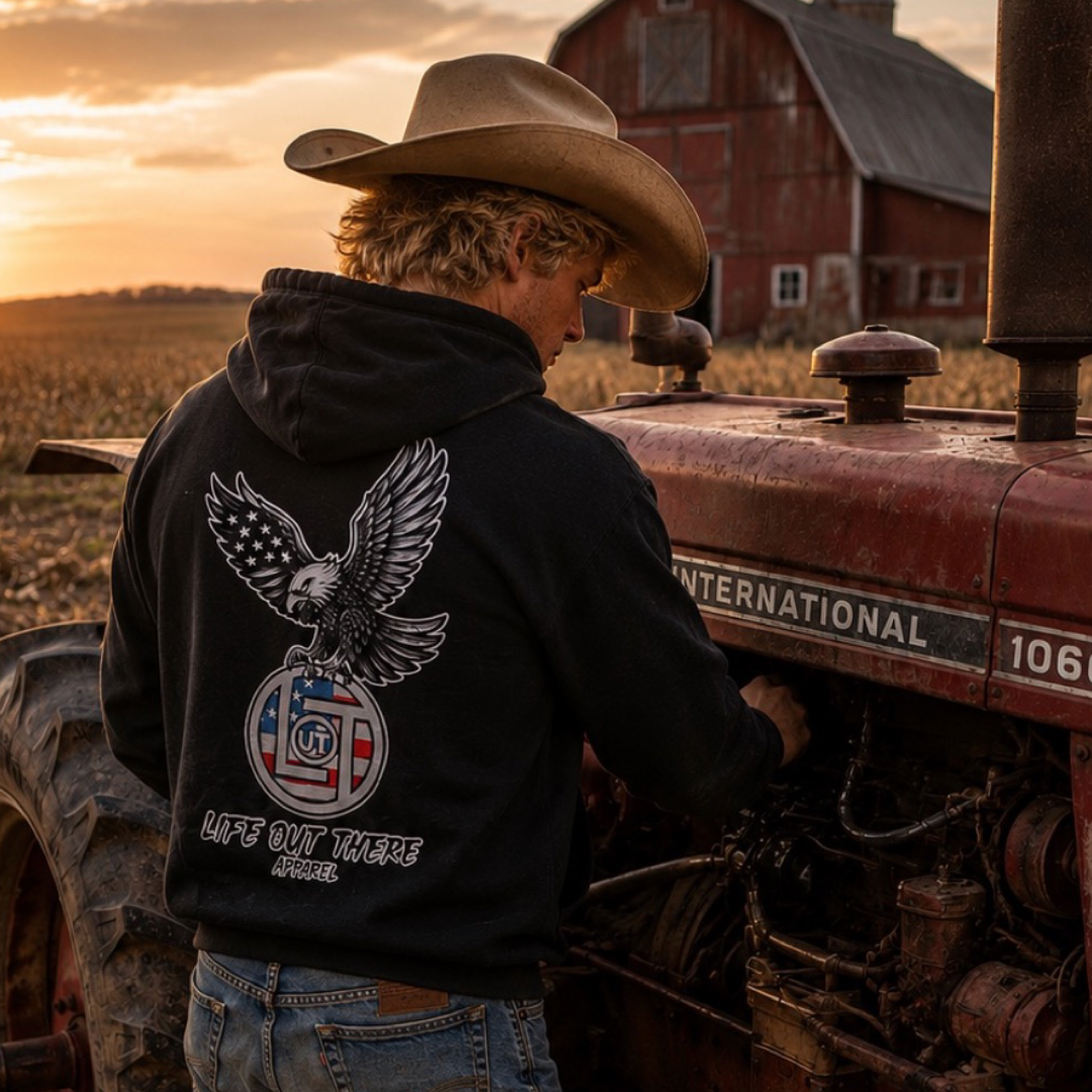 A man in a cowboy hat stands by a red tractor at sunset, with a barn behind him. He wears the Flight of Freedom Hoodie - Black by Life Out There Apparel, which features a soaring eagle graphic and brand text on the back.