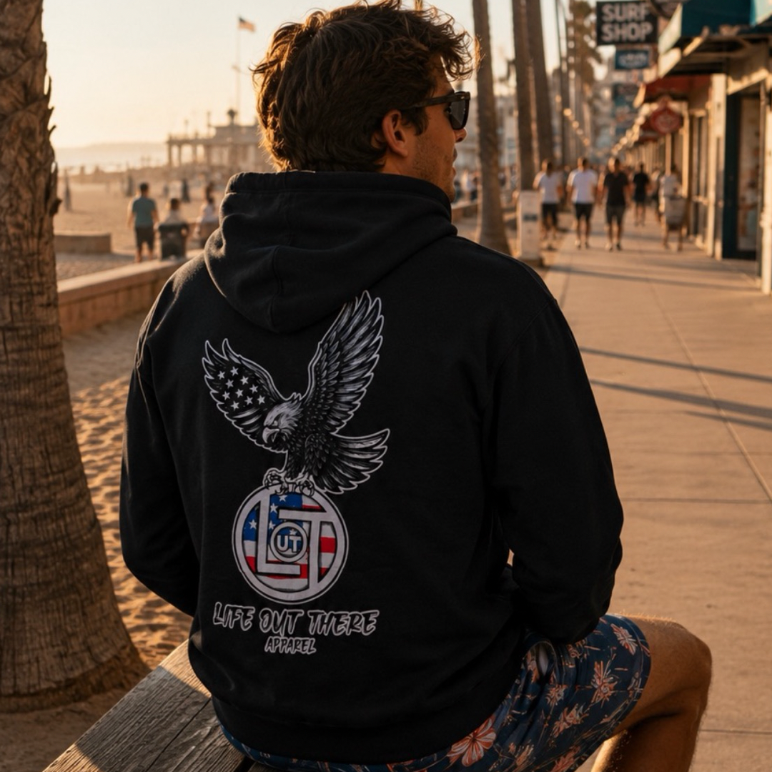 A man in sunglasses relaxes on a beach bench, sporting the Flight of Freedom Hoodie - Black from Life Out There Apparel, featuring an eagle graphic and "FLIGHT of FREEDOM" on the back. Sunlit boardwalk and palm trees complete the adventurous vibe.