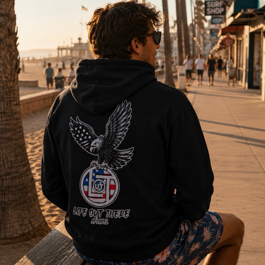 A man in sunglasses relaxes on a beach bench, sporting the Flight of Freedom Hoodie - Black from Life Out There Apparel, featuring an eagle graphic and "FLIGHT of FREEDOM" on the back. Sunlit boardwalk and palm trees complete the adventurous vibe.