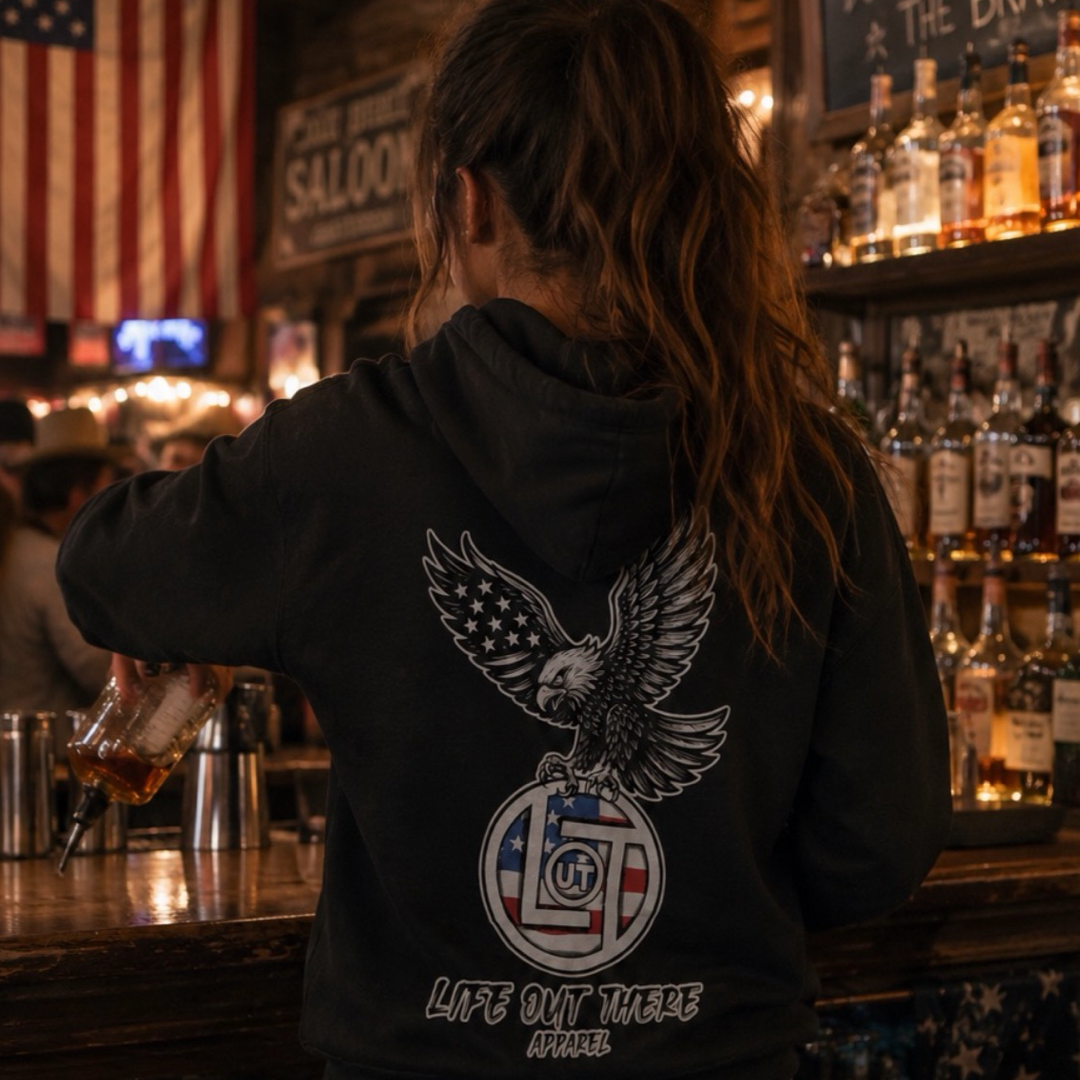 A person with long hair in a ponytail, wearing the Life Out There Apparel Flight of Freedom Hoodie - Black with an eagle and American flag, pours a drink behind a dimly lit bar decorated with liquor bottles and a U.S. flag.
