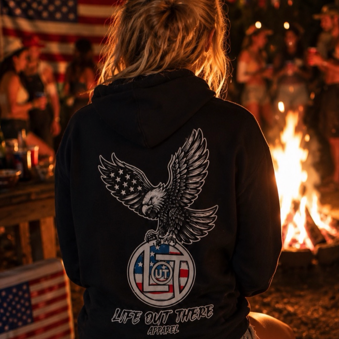 By a campfire at night, someone in a Life Out There Apparel Flight of Freedom Hoodie – Black sits near an American flag, surrounded by friends, capturing the adventurous spirit of this American anniversary apparel.