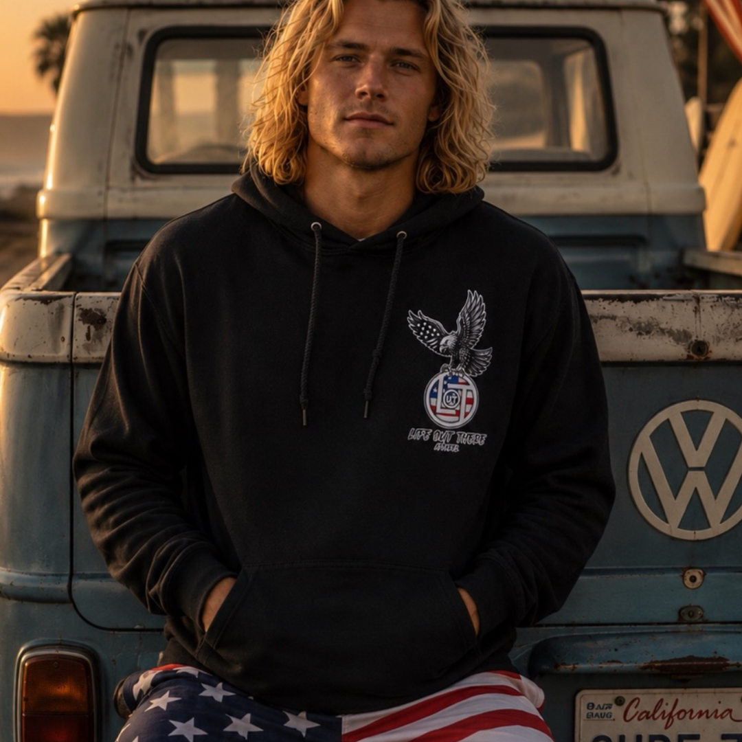 A young man with long blond hair sits on a vintage Volkswagen truck, wearing Life Out There Apparel’s Flight of Freedom Hoodie - Black and American flag shorts—a relaxed, beachy California vibe.