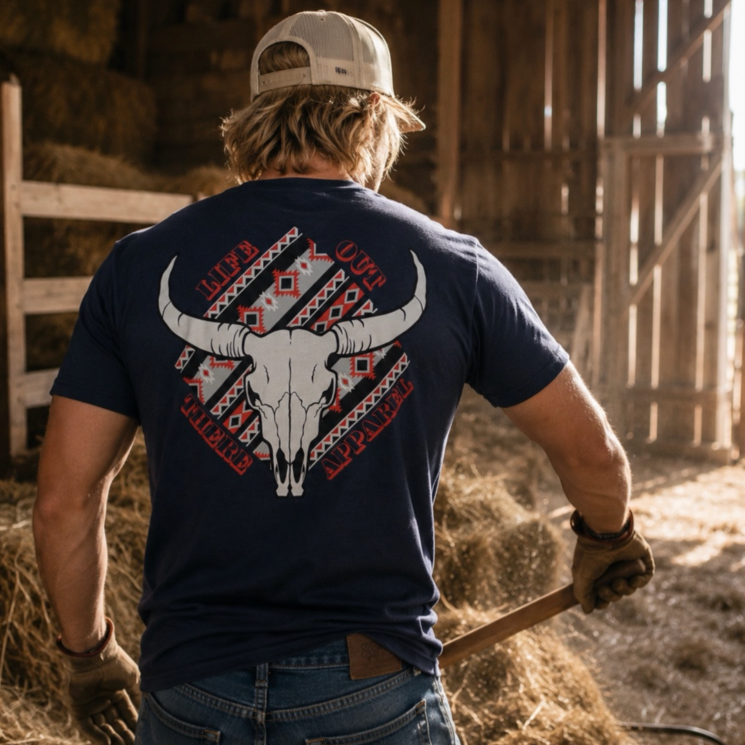 A person stands in a barn among hay bales, holding a tool and wearing the Longhorn Outlaw Tee - Navy from Life Out There Apparel, a unisex shirt featuring a bold bull skull graphic and LONGHORN OUTLAW text that combines rugged style with everyday comfort.