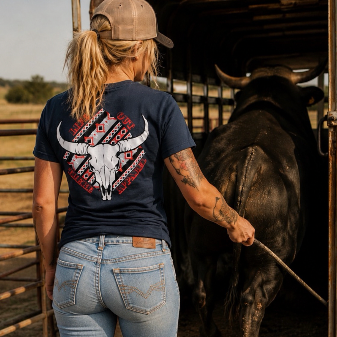 A person in a cap, jeans, and the Longhorn Outlaw Tee in navy from Life Out There Apparel—a unisex shirt—stands beside a large LONGHORN OUTLAW bull in a metal pen, holding a stick and facing away from the camera on a sunny day.