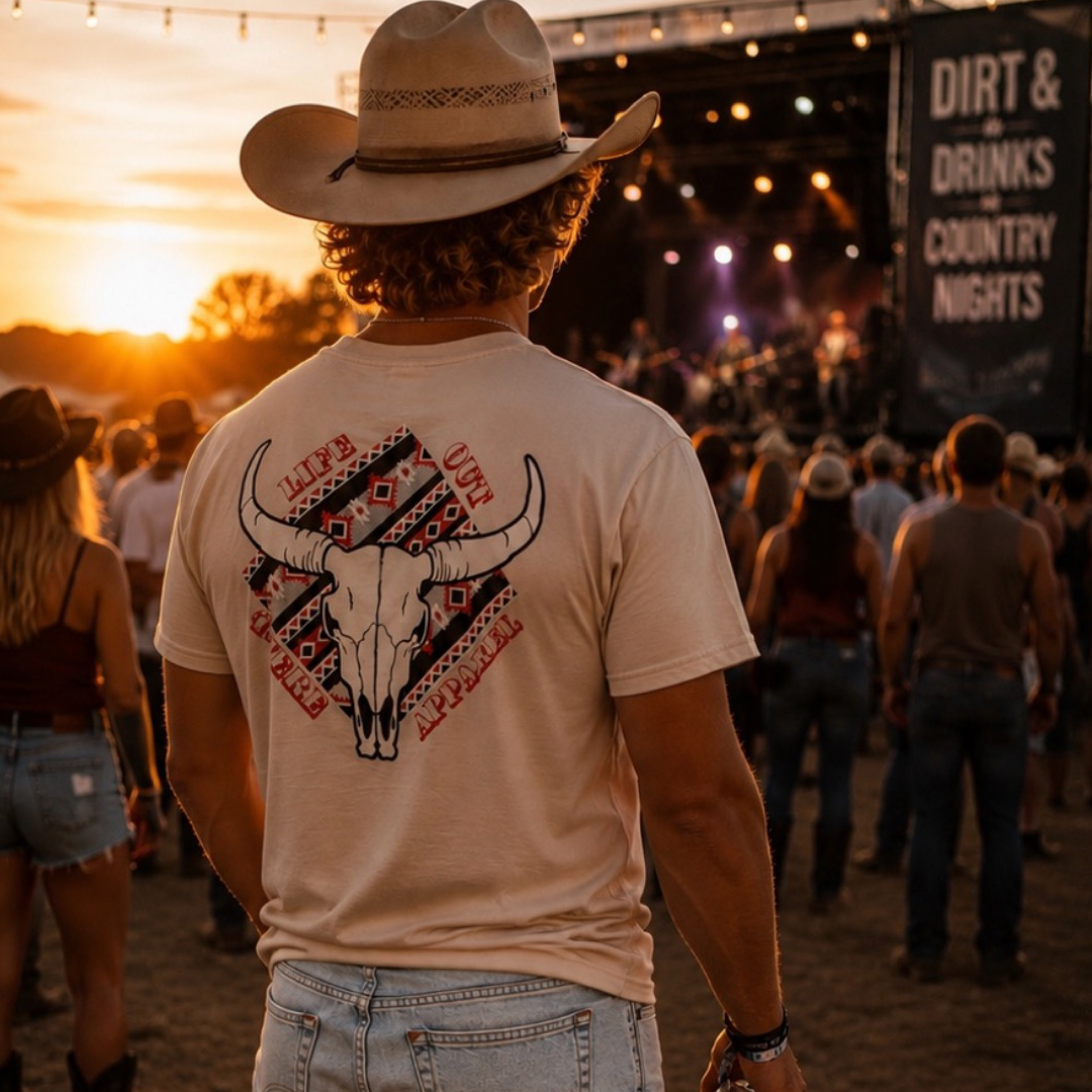 Wearing the Life Out There Apparel Longhorn Outlaw Tee in Sandstone and a cowboy hat, a man stands at an outdoor country music concert at sunset, facing the stage with festival lights and a lively crowd behind him.