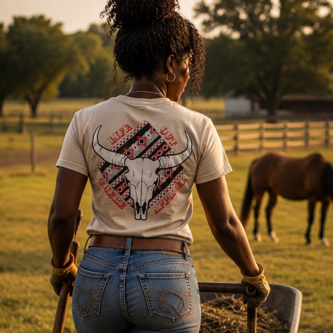 Wearing gloves and the Longhorn Outlaw Tee in Sandstone by Life Out There Apparel, a woman pushes a wheelbarrow through a grassy field, with a horse and trees visible in the background.