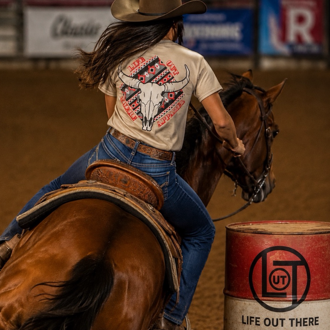 A woman in a cowboy hat rides a horse around a barrel in an arena. She wears jeans and the Longhorn Outlaw Tee - Sandstone—a stylish unisex shirt from Life Out There Apparel—featuring a steer skull graphic. The Life Out There Apparel logo appears in the lower right corner.