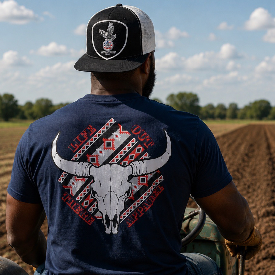 A man wearing the Longhorn Outlaw Tee in navy by Life Out There Apparel—a stylish unisex shirt—paired with a black-and-white trucker hat sits on a tractor, gazing out over freshly plowed fields beneath a blue, cloud-streaked sky.
