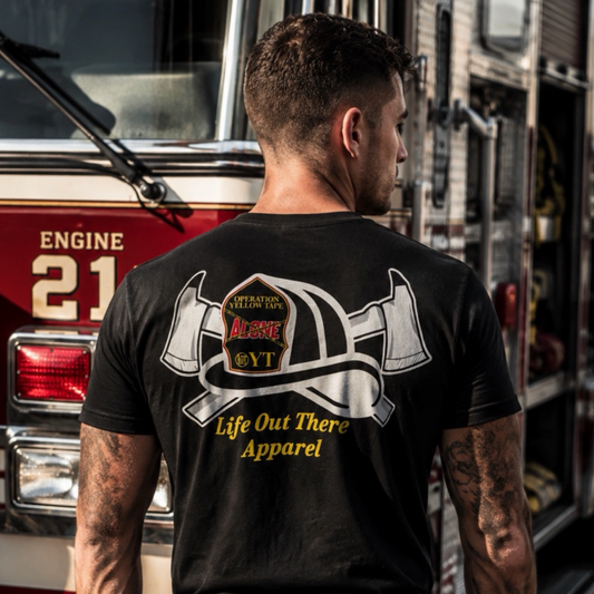 A man stands in front of a fire truck, wearing the Life Out There Apparel Drop The Shield Tee in black, featuring crossed axes, a helmet graphic, and text on the back—a powerful nod to suicide awareness within the firefighting community.
