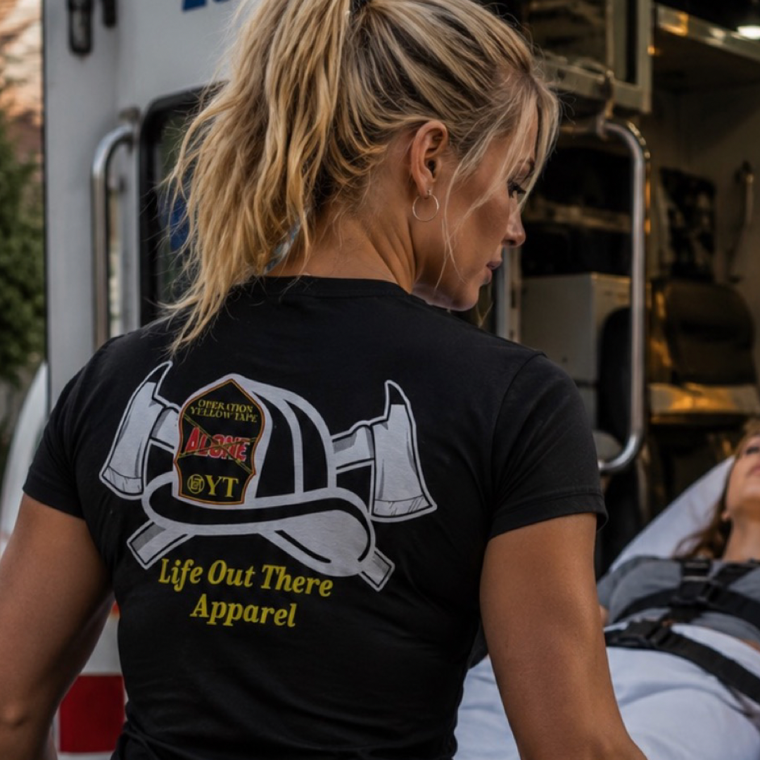 A blonde woman in a Life Out There Apparel Drop The Shield Tee – Black stands by a person on a stretcher near an ambulance, highlighting the brand’s commitment to suicide awareness and support during emergency situations.