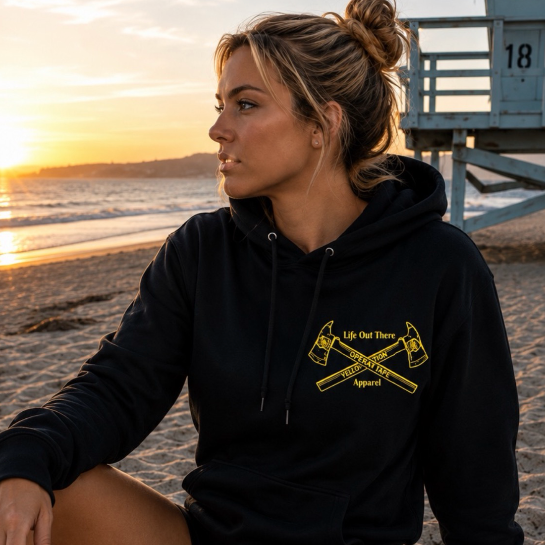 A woman sits on the beach at sunset wearing the Drop The Shield Hoodie in black from Life Out There Apparel. The hoodie features bold yellow text and two crossed axes, symbolizing support for suicide awareness. Behind her, a lifeguard tower stands along the tranquil shoreline.