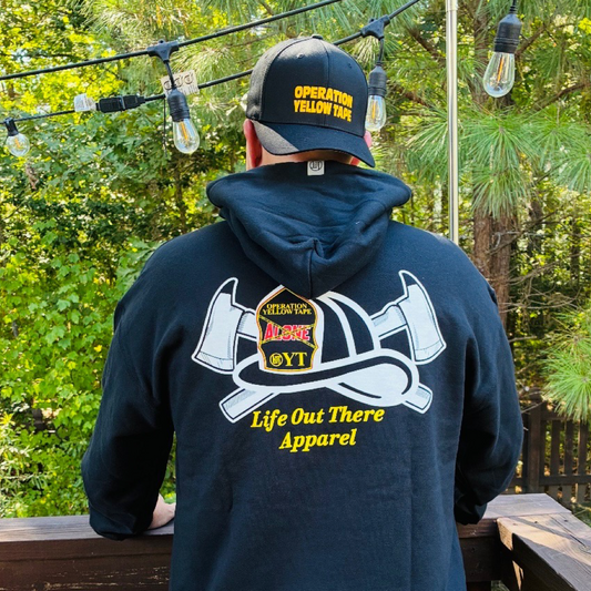 Wearing the black Drop The Shield Hoodie by Life Out There Apparel, a person stands outdoors among trees and string lights, raising suicide awareness in support of those who serve.