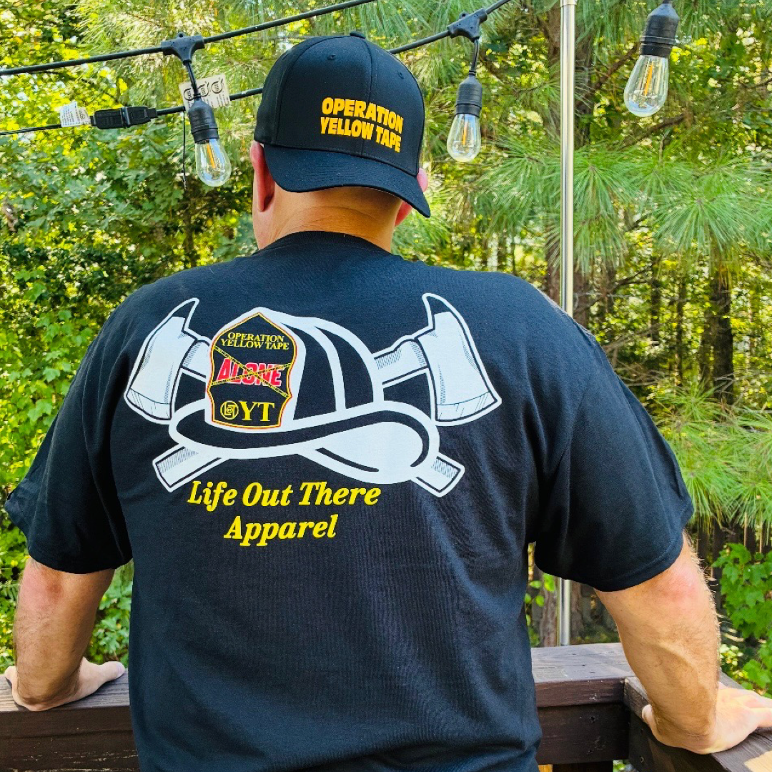 A man outdoors wears the Life Out There Apparel Drop The Shield Tee in black, which features a fire helmet and axes graphic along with the text “Operation Yellow Tape”—a tribute to suicide awareness. String lights and green trees are visible in the background.