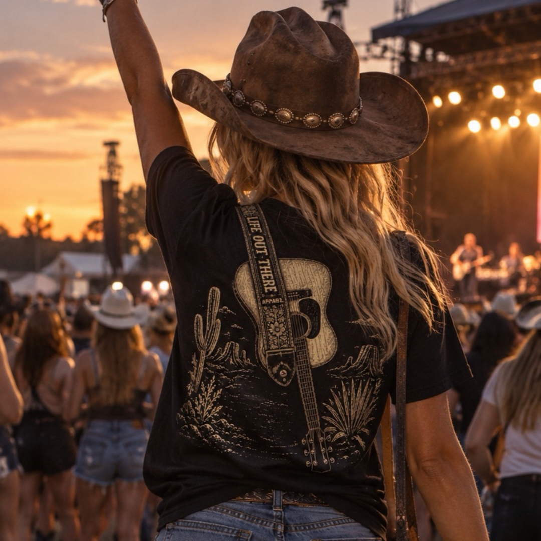 At sunset, someone in a cowboy hat and the Category Hurricane Tee - Black by Life Out There Apparel raises their arm at an outdoor concert, surrounded by a lively crowd and radiating true country style.
