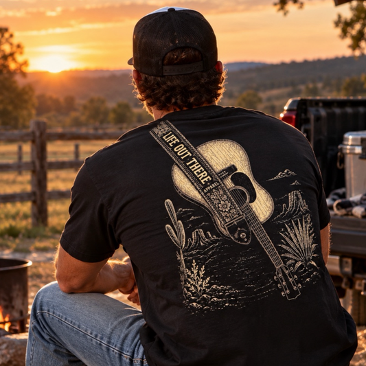 A man wearing the Category Hurricane Tee - Black by Life Out There Apparel sits by a campfire at sunset, facing a rural landscape with trees and a fence in the background.