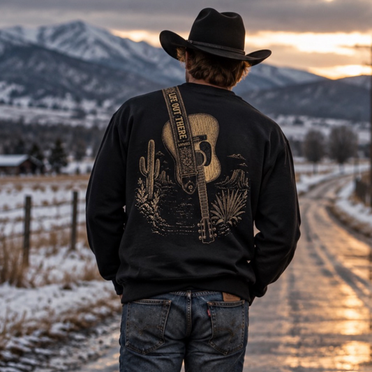 A person in a cowboy hat and jeans stands on a snowy rural road at sunset, mountains behind them. They wear the Category Hurricane Crewneck Sweatshirt - Black by Life Out There Apparel, capturing that true country vibe.