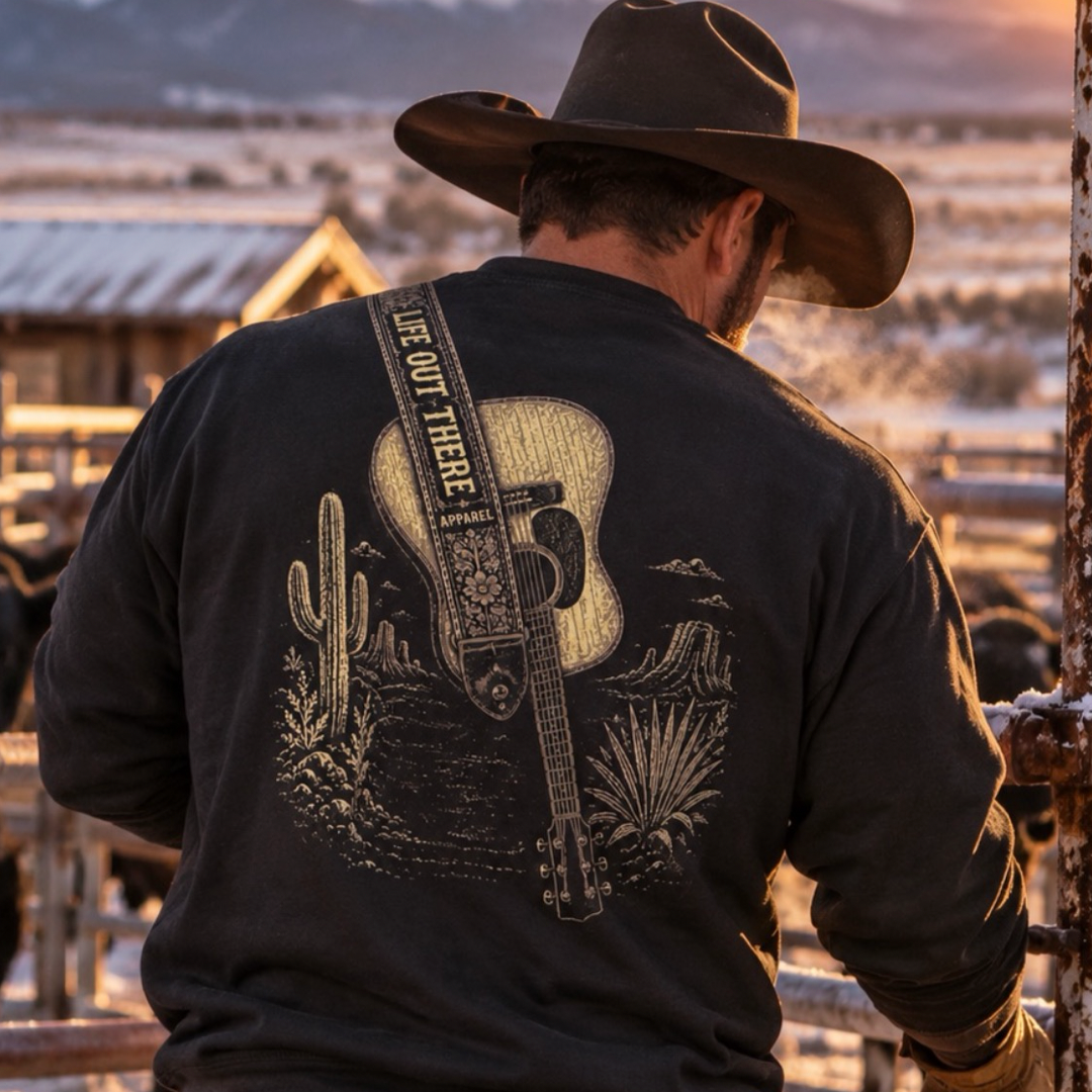 A man in a cowboy hat stands outdoors with a rustic vibe, wearing the Category Hurricane Crewneck Sweatshirt - Black by Life Out There Apparel, featuring a guitar, cactus, and desert design amid fences and cattle.